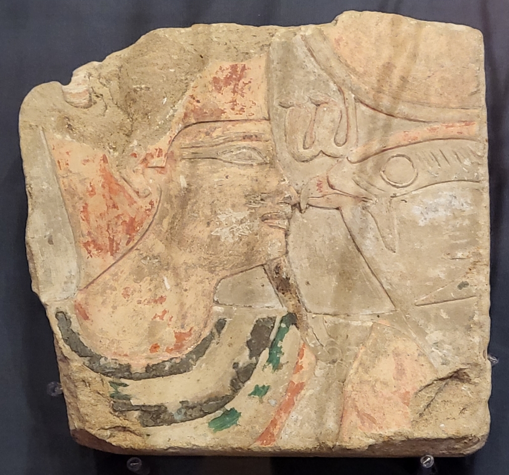 Image of a Pharaoh wearing the red crown and a coloured collar of black, green and red, nose to nose with a falcon god wearing a sun disc with a double uraeus.