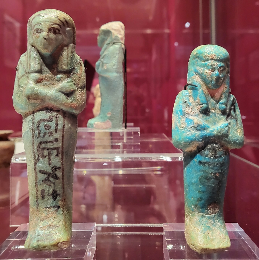 Two faience shabtis, with black ink inscriptions on their mummiform bodies, standing up in the case. 