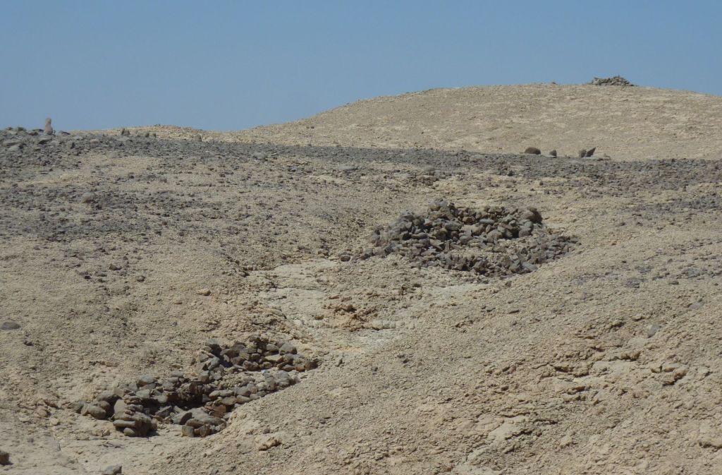 Part of the Hatnub desert. In the distance is a low hill crowned by a cairn, below that is a darker stony surface with several larger stones set upright, in the foreground is a shallow valley containing two habitation structures with various rooms.