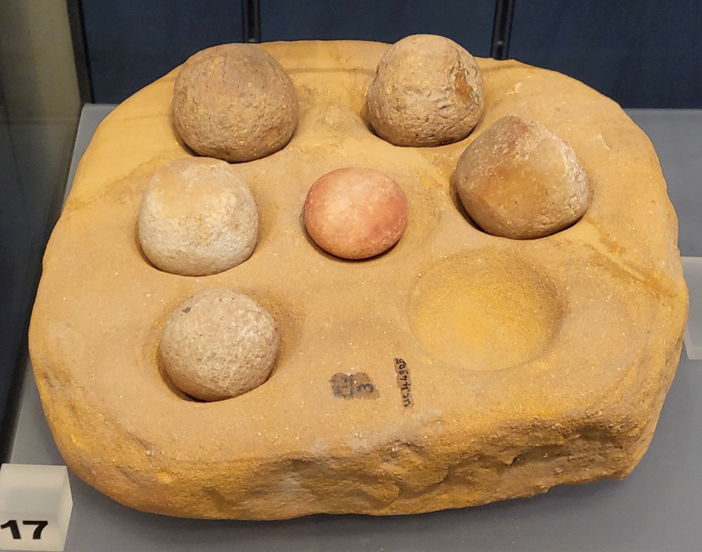 Picture of a stone block with seven rounded cups in the top. Various roughly rounded stones are present in six of the cups.