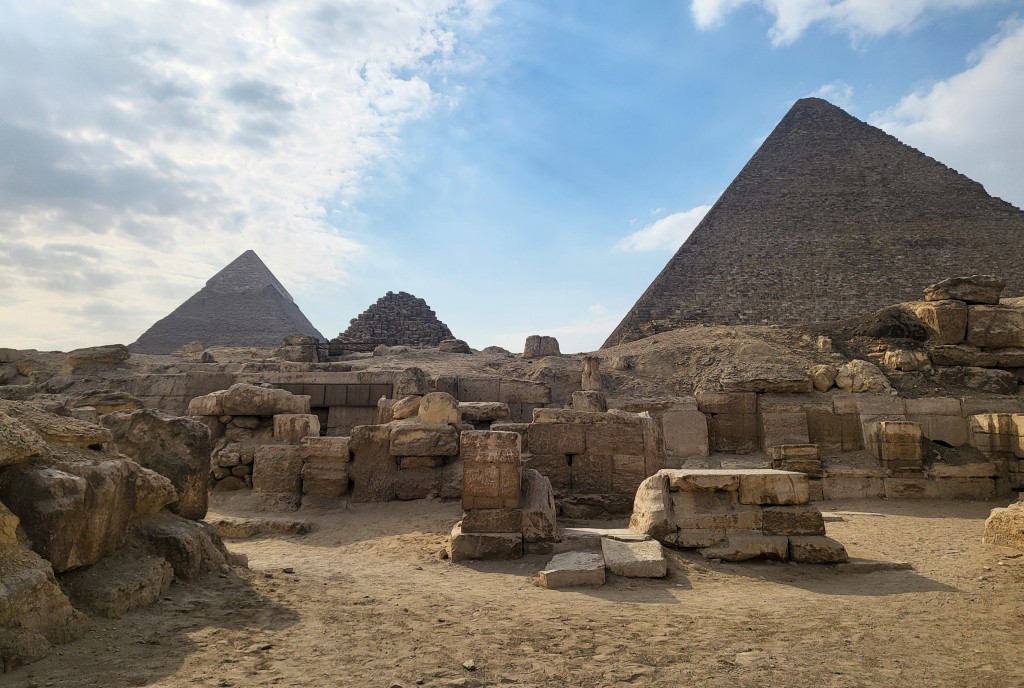 Photograph of large masonry-edged mastabas with pyramids in the background. A small chapel, built of cut blocks is visible in the centre in the 'street' between the mastabas.