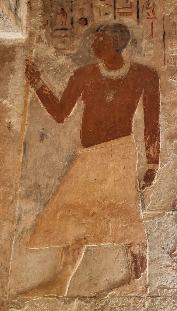 A relief of a portly man, with a short black wig, decorated collar and long kilt. He holds a long staff in his right hand and wears gold bracelets.