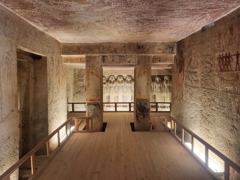 View of carved tomb chamber with decorated walls and a door cut into the left wall. Immediately ahead is a door and two side passages leading into another chamber with a row of statues carved out of the rear wall. Painted reliefs cover the walls.