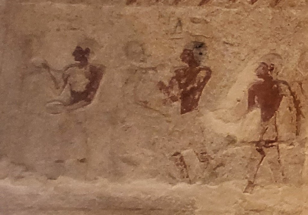 Detail of a scene showing three men, making funerary statues. The left man paints a standing statue. The centre man works on a seated statue and the right man carries something. The names of the first two men are written above their heads. 