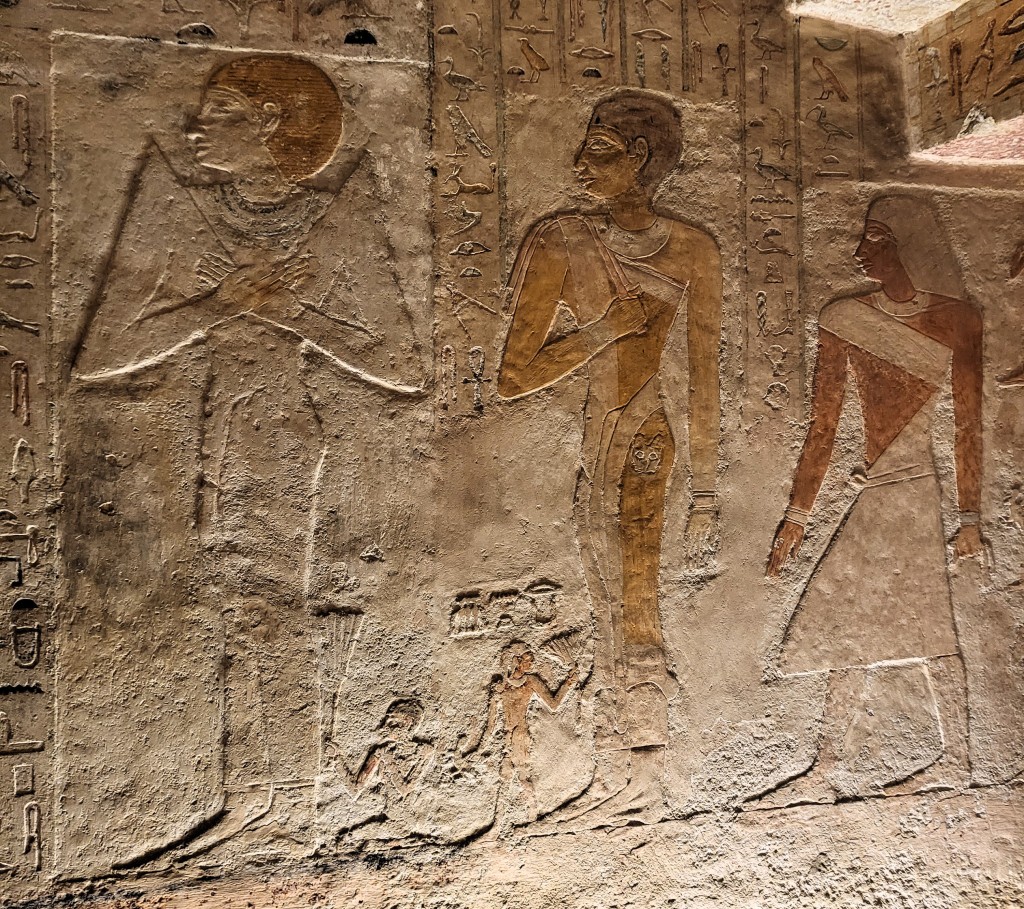 Photograph of a relief from Meresankh III's tomb, showing a woman in a right dress with pointed shoulders and a short red wig to the left; a woman in a sheath-type dress and a short black wig (centre); and a man, dressed as a lector priest with a pointed kilt and cross-body sash and a longer wig (right). 