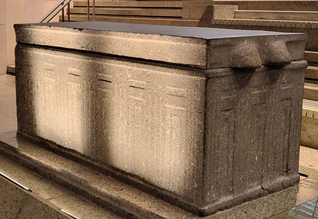 Photograph of a black granite  rectangular sarcophagus with lug handles at each end of the lid, decorated with recessed palace-facade panelling, with an inscription around the top and down one corner.